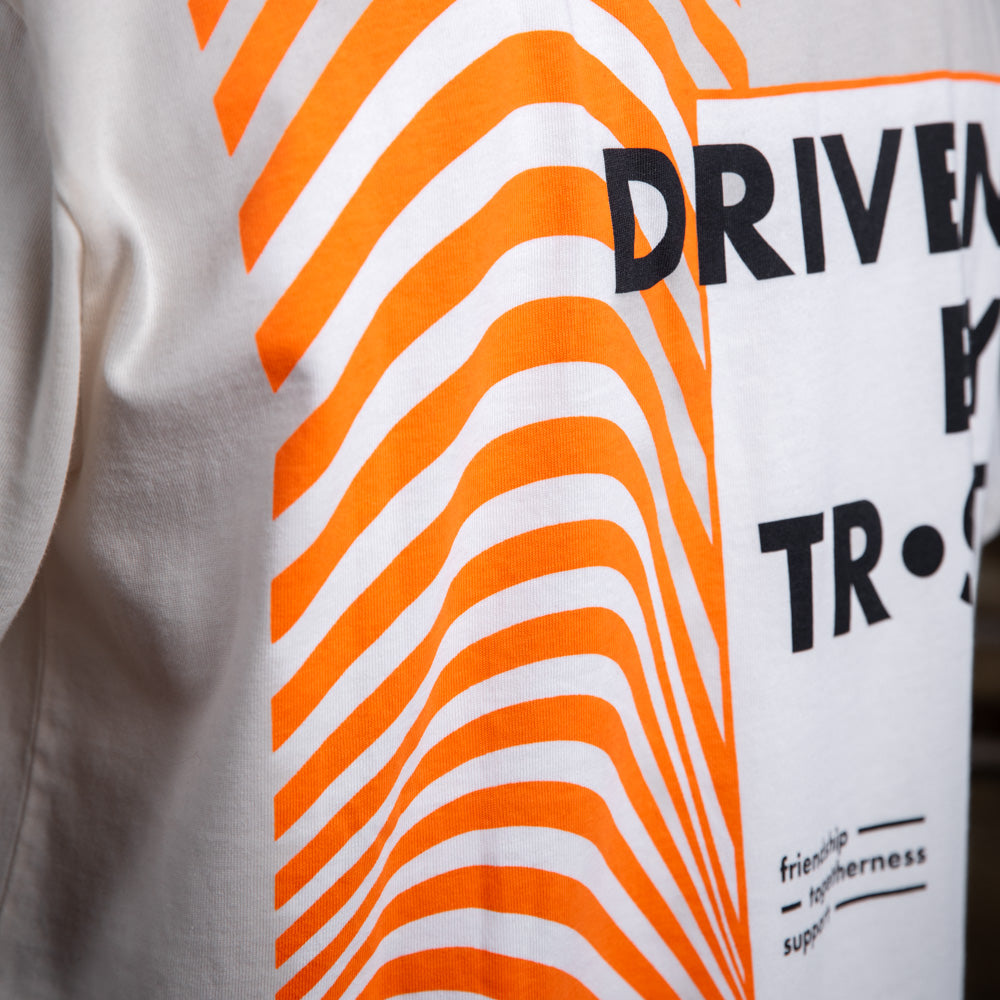 Nahaufnahme des Rückendrucks auf dem WE•TRST-T-Shirt „Driven by Trust“ mit leuchtend orangefarbenen Wellenmustern und Typografie.
