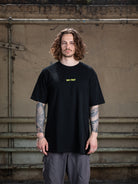 A male model with wavy brown hair and tattoos on both arms wearing a black WE•TRST oversized t-shirt with a yellow chest logo, standing in front of an industrial wall.