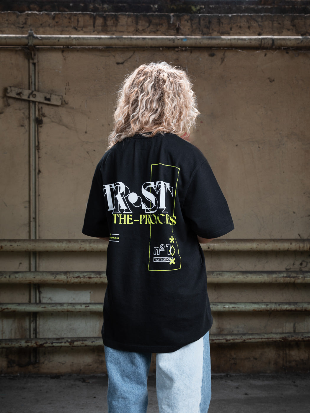 Back view of a female model with blonde curly hair wearing a black WE•TRST t-shirt featuring the large "TRST THE PROCESS" graphic print in white and yellow.