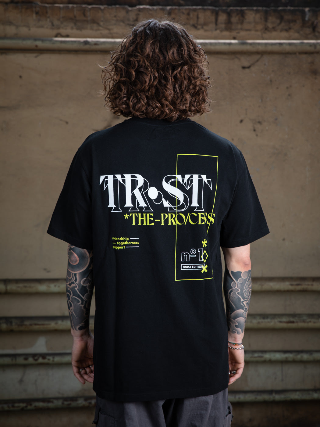 Close-up of the back design of a black WE•TRST t-shirt, showcasing the "TRST THE PROCESS" typography and graphic elements in white and neon yellow.