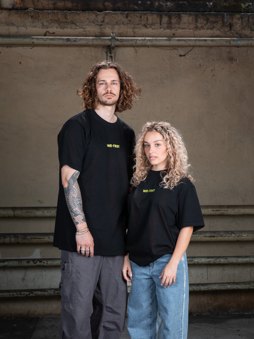 A male model in a black WE•TRST t-shirt with tattoos on his arms, paired with cargo pants, standing against a raw concrete wall.