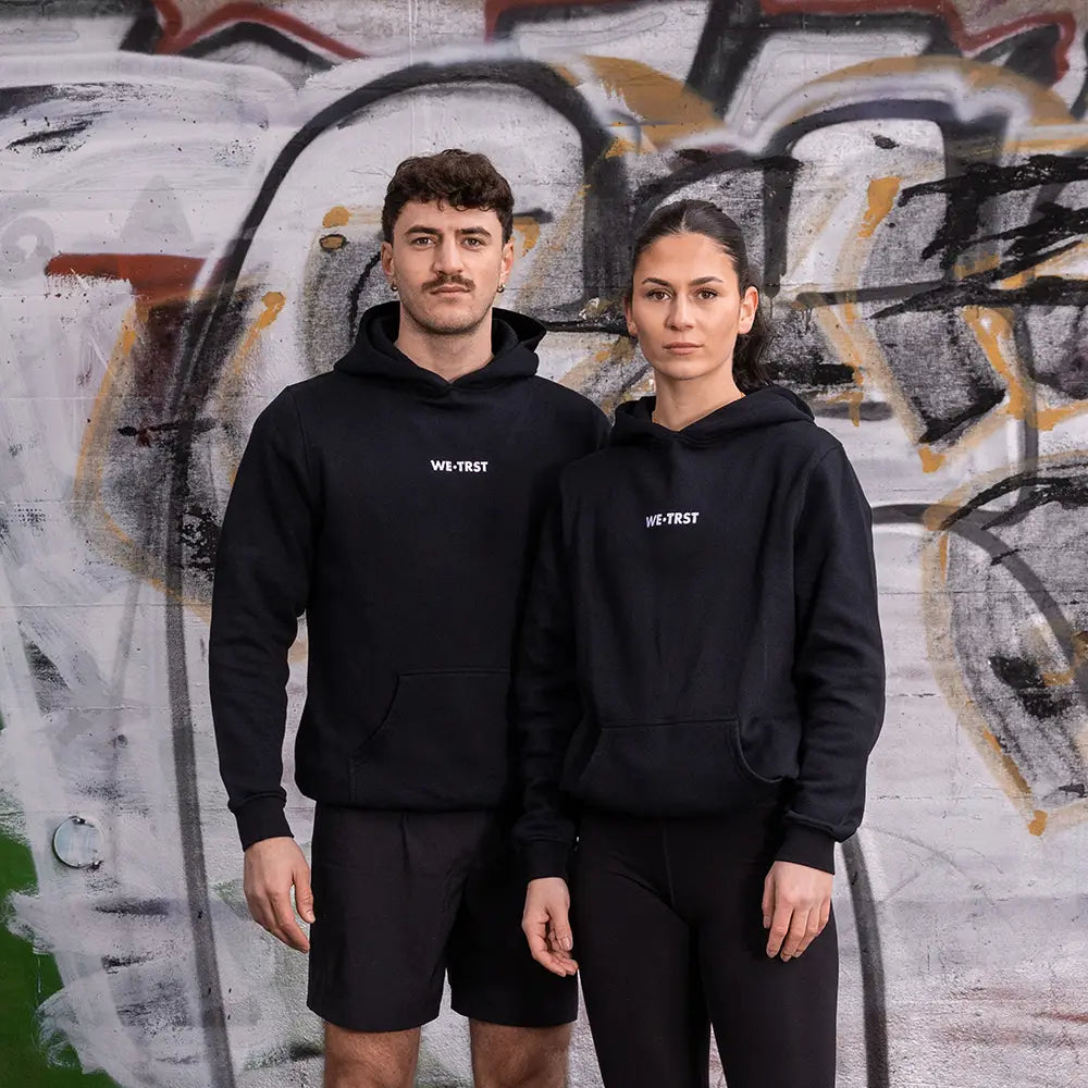 WE•TRST Running Department Hoodie – schwarze Unisex Streetwear für Läufer:innen vor urbaner Graffiti-Wand
