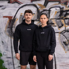 WE•TRST Running Department Hoodie – schwarze Unisex Streetwear für Läufer:innen vor urbaner Graffiti-Wand