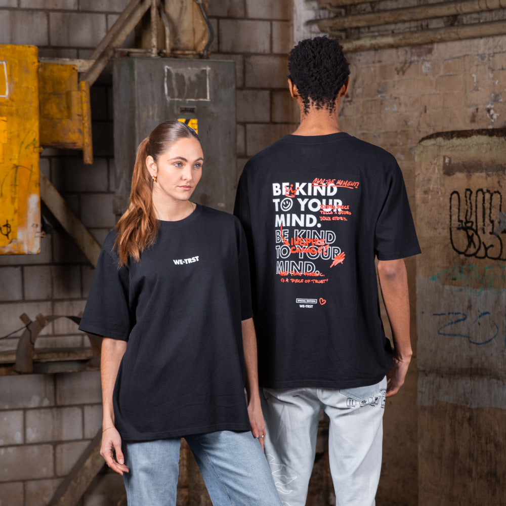 Zwei Models, die schwarze WE•TRST-T-Shirts in Übergröße mit einem Motiv für psychische Gesundheit tragen, posieren in einer städtischen Industriekulisse mit Graffiti-verschmierten Wänden.