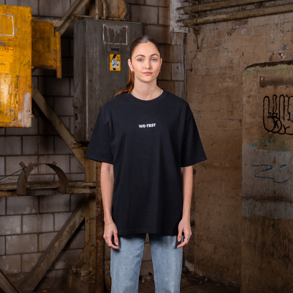 Weibliches Modell, das ein schwarzes WE•TRST-T-Shirt in Übergröße mit minimalistischem Logo trägt, posiert in einer städtischen Industrieumgebung mit freiliegenden Ziegelwänden und Metallstrukturen.