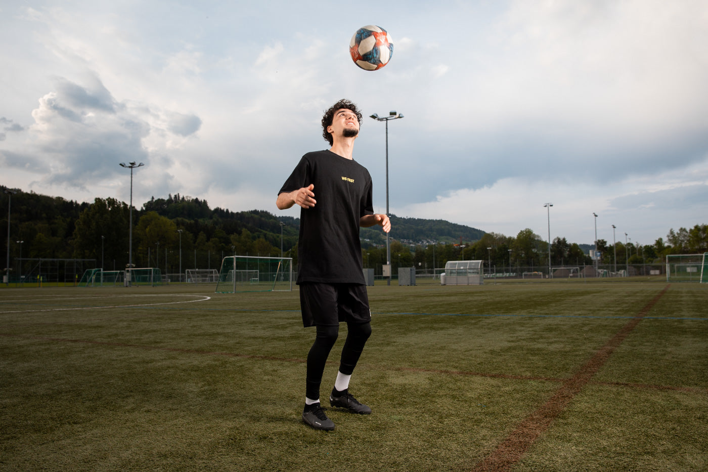 in Mann spielt mit Leidenschaft Fußball auf dem grünen Rasen. Er trägt ein dynamisches T-Shirt von WE•TRST, das seinen Einsatz für Zusammenarbeit und Vertrauen verdeutlicht.